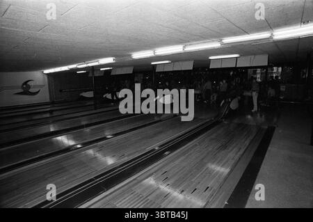 Compétition de bowling., Whizgle News, Dutch Desk, pays-Bas, 1950 - 2000. Voici les rubriques de l'image. La scène capture une piste de bowling, caractérisée par ses longues pistes en bois poli s'étendant au loin. La surface brillante reflète l'éclairage aérien, qui projette une lueur douce et silencieuse dans tout l'espace. Les couloirs sont flanqués de bancs bas et rembourrés où des groupes de personnes se rassemblent, certains engagés dans la conversation tandis que d'autres se concentrent sur le jeu à portée de main. Vers la fin des voies, une rangée de pare-chocs dépasse, peint en blanc avec des accents noirs, suggérant un élément Banque D'Images