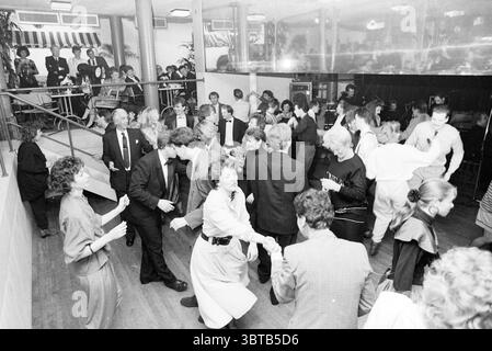 Festiball Concertgebouw Football Haarlem, Whizgle News, Dutch Desk, pays-Bas, 1950 - 2000 le 28-01-1989. L'image contient ces rubriques. La scène est animée et remplie de mouvement, représentant un rassemblement social bondé, probablement une danse ou une célébration. Au premier plan, un groupe diversifié de personnes est engagé dans la danse, échangeant des sourires et des rires. La tenue est formelle, avec de nombreuses personnes vêtues de costumes et de robes élégantes, présentant un mélange de couleurs sombres et claires, ainsi que des motifs tels que des rayures et des fleurs. Certaines tenues ont un style des années 80, avec des épaulettes et des styles audacieux creat Banque D'Images