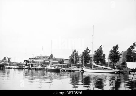 Voile dans la région de Haarlem voile, Whizgle News, Dutch Desk, pays-Bas, 1950 - 2000 le 28-07-1964. Ces rubriques sont illustrées dans l'image. La scène présente un port tranquille, caractérisé par une surface d'eau calme reflétant les bateaux et les bâtiments environnants. De nombreux navires sont amarrés à la marina en bois, variant en taille et en conception, certains semblant être des voiliers tandis que d'autres sont des bateaux à moteur. Les couleurs sont atténuées, dominées par des nuances de gris et de blanc, avec des contrastes doux indiquant un temps couvert. Les bâtiments bordant le port sont modernes, construits à partir de matériaux tels que Banque D'Images