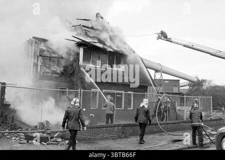 Lutte contre l'incendie à la maison de démolition Velsen-Noord (à côté de la centrale électrique) Velsen-Noord, Whizgle News, Dutch Desk, pays-Bas, 1950 - 2000 on 30-10-1980. L'image contient ces rubriques. La scène est remplie d'un sentiment d'urgence et de chaos alors que de la fumée épaisse et tourbillonnante s'écoule d'un bâtiment partiellement effondré, indiquant un incendie important. La structure, une maison de deux étages avec un toit de chaume, montre des signes de graves dommages ; des sections du toit se sont effondrées, et des flammes peuvent être vues lécher vers le haut de l'intérieur. Les pompiers portant des casques et des équipements distinctifs sont activement engagés dans la lutte contre le bla Banque D'Images