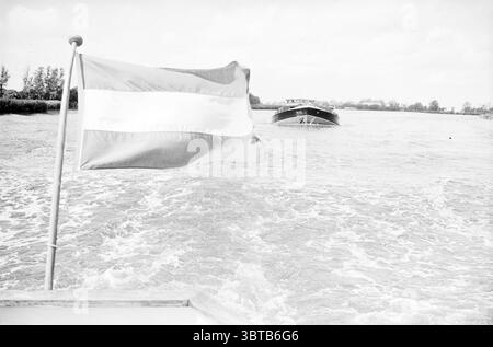Bateau d'excursion à Groenendaal avec chauffeur Alderman, Whizgle News, Dutch Desk, pays-Bas, 1950 - 2000 le 28-06-1962. Ce sont les éléments de l'image. La scène représente une voie navigable bordée de verdure luxuriante de chaque côté. Un drapeau, bien visible au premier plan, présente des bandes horizontales audacieuses de rouge, blanc et vert. Le tissu du drapeau bat doucement dans la brise, introduisant une sensation de mouvement. En arrière-plan, un bateau navigue dans l'eau, créant des ondulations qui s'étendent vers l'extérieur, capturant l'essence dynamique de la scène. L'eau elle-même semble réfléchissante, s'affichant Banque D'Images