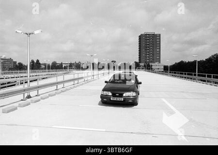 Ouverture du parking Schalkwijk Haarlem pays-Bas, Whizgle News, Dutch Desk, pays-Bas, 1950 - 2000 le 19-07-1991. Ces rubriques sont illustrées dans l'image. La scène capture une structure de stationnement ouverte et dure qui s'étend au loin. Dominant le premier plan est une berline de couleur foncée garée bien en vue, face au spectateur. Les lignes épurées de la voiture contrastent avec la surface grise en béton, qui s'étend vers l'extérieur et est marquée par des lignes de stationnement blanches et faibles. En arrière-plan, un grand bâtiment rectangulaire se dessine avec une série de fenêtres reflétant l'atmosphère silencieuse Banque D'Images