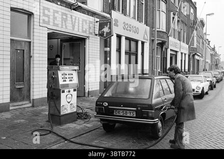 Garage avec pompe à essence, Whizgle News, Dutch Desk, pays-Bas, 1950 - 2000 sur 01-1980. Ces rubriques apparaissent dans l'image. Dans un cadre urbain animé, la scène capture un moment d'une époque révolue. Le point focal est une station-service vintage, caractérisée par son extérieur carrelé, avec une grille de carreaux pâles, blanc cassé et légèrement usés. Au-dessus de la zone de service, le mot « SERVICE » est bien en évidence, bien que les lettres soient partiellement masquées, ajoutant une impression d'âge à l'environnement. À gauche, un homme vêtu d'un manteau noir et d'un chapeau se tient à une aire de service ouverte, engagé dans une activité, comme Banque D'Images
