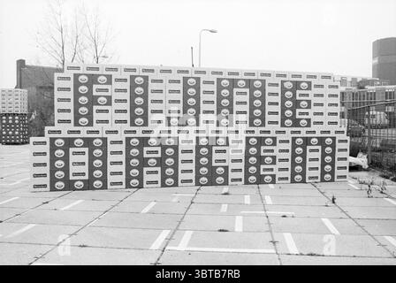 Caisses de la société Rieu : Happy Days Christmas and Christmas Trees, Whizgle News, Dutch Desk, pays-Bas, 1950 - 2000 sur 15-12-1987. Ce sont les éléments de l'image. Dans cette scène, une série de boîtes empilées crée un arrangement visuel saisissant sur un fond industriel. Les boîtes sont principalement blanches avec diverses nuances de gris et comportent des logos circulaires de couleurs vives, suggérant probablement une marque ou un produit. Les motifs répétitifs des logos contribuent à une qualité rythmique dans la composition. Le cadre semble être une zone urbaine, avec la chaussée en béton partiellement v Banque D'Images