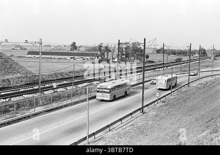 Train et bus Haarlem - Amsterdam Noord Zuid Hollandse Vervoersmaatschappij N.V. NZ, Whizgle News, Dutch Desk, pays-Bas, 1950 - 2000 on 11-06-1964. L'image inclut ces rubriques. La scène capture une route très fréquentée le long d'une voie ferrée. Sur la route, deux bus sont proéminents, l’un se dirigeant vers le spectateur tandis que l’autre semble être dans une voie parallèle, tous deux rendus dans des nuances de gris subtiles. Les autobus possèdent des formes arrondies et de grandes fenêtres, ce qui fait allusion à leur conception fonctionnelle pour le transport en commun. Le cadre est caractérisé par un paysage tentaculaire qui s'étend dans la BA Banque D'Images