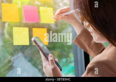 Jeune femme d'affaires asiatique brainstorming stratégie tenant le téléphone et écrivant de nouvelles idées sur le mur de verre avec des notes adhésives. Mise au point sélective. Banque D'Images