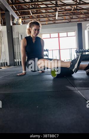 Femme portant des vêtements d'entraînement roulant mollet sur rouleau de mousse sur tapis en caoutchouc près de la fenêtre dans le gymnase Banque D'Images