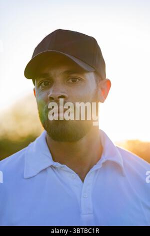 Homme dans les années 30 posant dehors au coucher du soleil portant une casquette de baseball noire et un polo blanc Banque D'Images