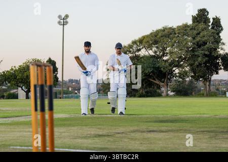 Divers coéquipiers de cricket marchant côte à côte sur le terrain près de souches de bois tenant des chauves-souris en bois Banque D'Images