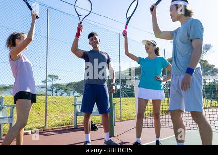 Divers amis élevant des raquettes en vêtements de sport et célébrant point sur le court de tennis rouge près du filet Banque D'Images