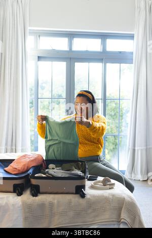 Femme asiatique à la fin des années 30 sur le lit dans la valise d'emballage de chambre examinant le dessus vert à côté des écouteurs Banque D'Images