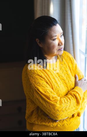 Femme asiatique senior en pull jaune debout à la maison par fenêtre croisant les bras regardant les rideaux Banque D'Images