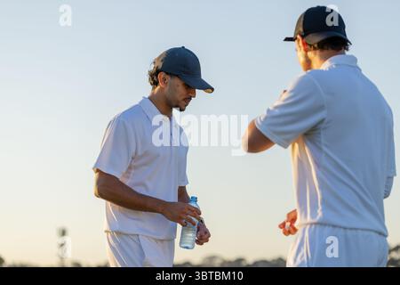 Divers coéquipiers masculins portant des polos blancs et des casquettes debout sur le terrain de cricket, tenant une bouteille d'eau Banque D'Images