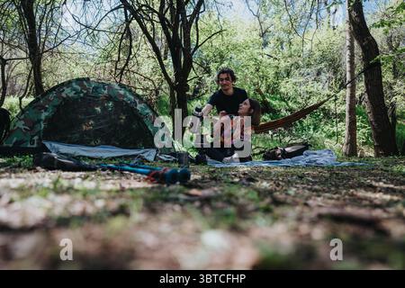 Jeune couple profitant d'une journée de camping en plein air dans une forêt, assis à côté d'une tente, entouré par la nature. Banque D'Images