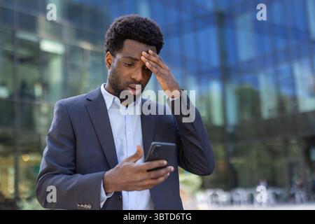 Homme d'affaires afro-américain anxieux regardant son téléphone en détresse devant un immeuble de bureaux moderne. Il semble préoccupé par ce qu'il voit à l'écran. Banque D'Images