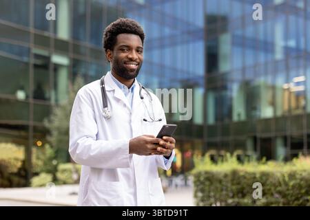 Un médecin africain en manteau blanc sourit tout en tenant un smartphone devant un bâtiment moderne. Banque D'Images