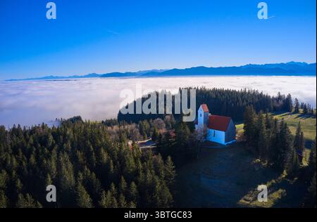 Vue aérienne de la chapelle blanche et dure avec son toit rouge se dressant contre la forêt vert profond, surplombant une mer de nuages, Bernbeuren, Bavière, Allemagne. Banque D'Images
