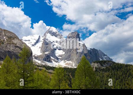 Les majestueux sommets enneigés de Marmolada s'élèvent bien au-dessus d'une forêt verdoyante, sous un ciel bleu vif avec des nuages blancs éparpillés. Banque D'Images