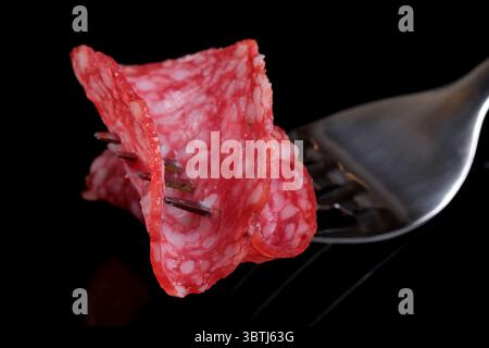 Délicieuse saucisse de salami parfumée finement tranchée sur une fourchette en acier gros plan sur un fond miroir noir Banque D'Images
