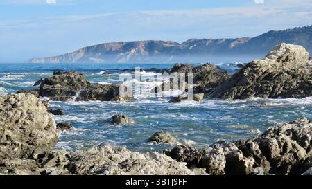 Paysage côtier accidenté avec des affleurements rocheux et des vagues écrasantes le long de la côte est de l'île du Sud de Nouvelle-Zélande, avec des montagnes brumeuses Banque D'Images