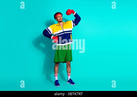 Jeune homme athlétique posant avec confiance dans des vêtements de sport rétro et des gants de boxe sur un fond sarcelle Banque D'Images