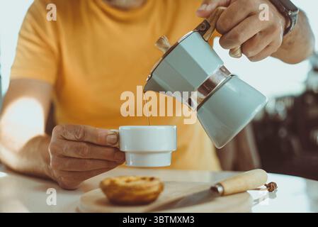 Homme assis à une table avec une pâtisserie pastel de nata versant une tasse de café d'une cafetière expresso italienne Banque D'Images