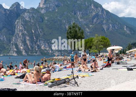 Nago-Torbole, Trentin, Italie - 15 juillet 2025 : les touristes apprécient la journée ensoleillée à Spiaggia Lungolago à Torbole sur la rive nord du lac de Garde. La plage de galets est très fréquentée, avec beaucoup de gens allongés sur des serviettes ou nageant dans l'eau claire *** Touristen genießen den sonnigen Tag am Spiaggia Lungolago à Torbole am nördlichen Gardaseeufer. Der Kiesstrand ist gut besucht, viele Menschen liegen auf Handtüchern oder baden im klaren Wasser Banque D'Images