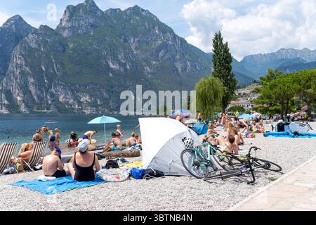 Nago-Torbole, Trentin, Italie - 15 juillet 2025 : les touristes apprécient la journée ensoleillée à Spiaggia Lungolago à Torbole sur la rive nord du lac de Garde. La plage de galets est très fréquentée, avec beaucoup de gens allongés sur des serviettes ou nageant dans l'eau claire *** Touristen genießen den sonnigen Tag am Spiaggia Lungolago à Torbole am nördlichen Gardaseeufer. Der Kiesstrand ist gut besucht, viele Menschen liegen auf Handtüchern oder baden im klaren Wasser Banque D'Images