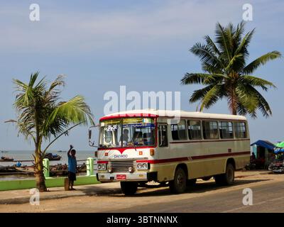 Bus garé au bord de la mer à Myeik, Myanmar, avec des palmiers, des bateaux, et une personne agitant dans un cadre côtier tropical animé sous un ciel clair. Banque D'Images