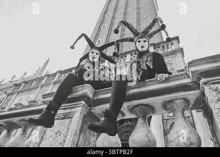 Deux personnages masqués en costumes de bouffon élaborés assis sur une balustrade de marbre pendant le Carnaval de Venise, avec le Campanile de Mark en arrière-plan Banque D'Images