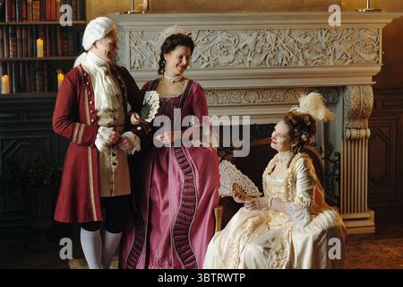 Homme d'âge moyen caucasien et deux femmes d'âge moyen caucasiennes portant des costumes d'époque baroque debout et assis près d'une cheminée ornée tenant des ventilateurs décoratifs et souriant Banque D'Images