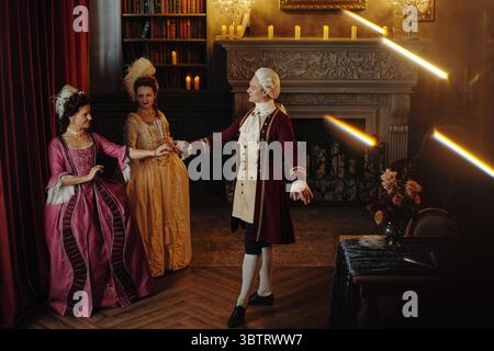 Homme d'âge moyen caucasien en costume baroque dirigeant deux jeunes femmes adultes caucasiennes en robes de style Renaissance, debout dans une bibliothèque ornée de bougies et d'un décor classique Banque D'Images