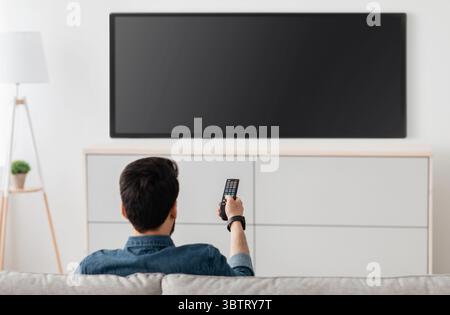 Homme regardant la télévision, allumant le téléviseur à écran plat plasma, pointant la télécommande vers un écran de télévision vide sur un mur blanc Banque D'Images