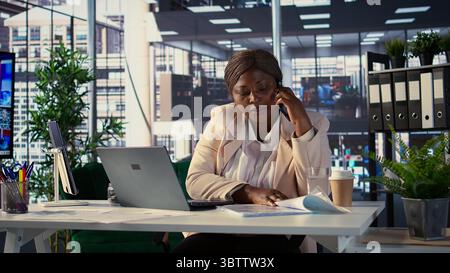 Une entrepreneure afro-américaine travaille sur la planification financière sur son téléphone portable, répondant à un appel des investisseurs pour partager des idées et des données. Femme PDG fait des tâches commerciales sur appel téléphonique. Caméra B. Banque D'Images