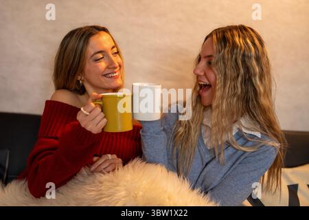 Deux amies joyeuses appréciant un moment de détente ensemble, grillant avec des tasses et partageant le rire dans un cadre confortable à la maison Banque D'Images