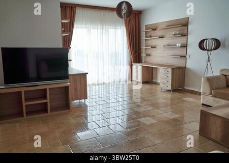 Un salon moderne avec une grande fenêtre avec lumière naturelle, carrelage brun, et mobilier en bois minimaliste, créant un espace spacieux et invi Banque D'Images