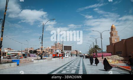 Les gens se promènent dans les rues animées de Kufa Irak, profitant d'une journée ensoleillée et de la culture locale, avec une belle mosquée en vue. Banque D'Images
