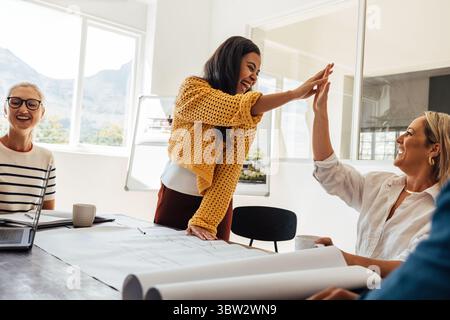 De jeunes femmes architectes collaborant dans un bureau lumineux, partageant des idées et célébrant le succès avec un high-Five, mettant en valeur le travail d'équipe, l'enthousiasme et Banque D'Images