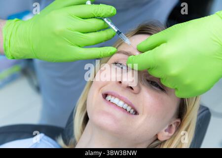 Une procédure esthétique professionnelle : gros plan d'une femme souriante recevant une injection de Botox dans une clinique moderne avec des gants verts et confortables Banque D'Images