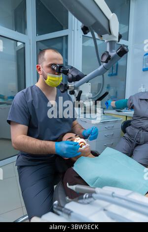 Dentiste effectuant une procédure sous microscope dans le cabinet dentaire moderne avec le patient détendu pour le traitement et assistant sur les outils de préparation de fond Banque D'Images