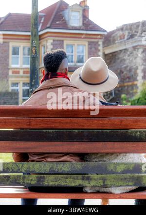 Un couple est assis sur un banc de parc, la tête de l'homme reposant sur l'épaule de la femme. L'homme porte une veste en cuir marron et une écharpe rouge, w Banque D'Images