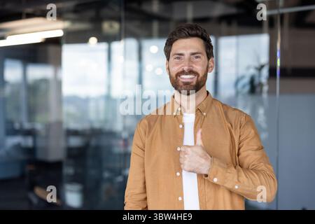 Un homme souriant et barbu donne un coup de pouce dans un cadre de bureau moderne, exsudant positivité et approbation. Banque D'Images
