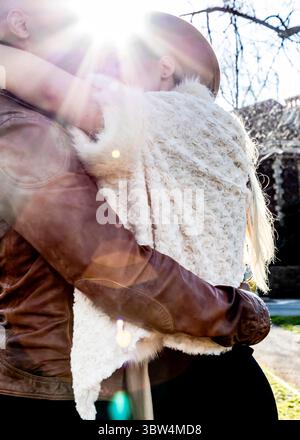 Un couple s'embrasse, l'homme portant une veste en cuir marron et la femme portant un gilet de fourrure blanche et un chapeau. Le soleil brille brillamment derrière eux. Banque D'Images