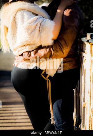 Une femme enceinte est embrassée par une personne portant une veste en cuir marron. Ils se tiennent près d'une structure métallique rouillée. Banque D'Images