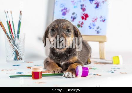Chiot Mongrel avec des pinceaux, des peintures et un chevalet avec une image sur fond clair Banque D'Images