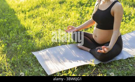 Gros plan de la femme enceinte médite tout en étant assis sur un tapis sur la pelouse par une soirée d'été ensoleillée. Concept de pacification et boost énergétique. Banne Banque D'Images