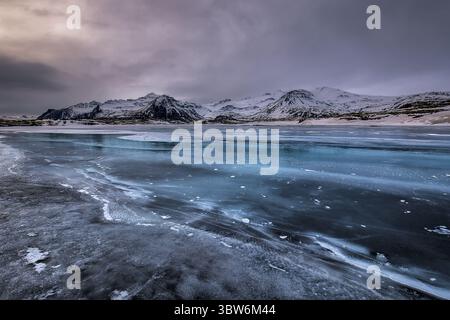 Vue de motifs glacés dansant sur le lac partiellement gelé, reflétant les montagnes enneigées sous un ciel texturé, Vik, Islande. Banque D'Images