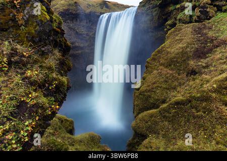 Vue sur la cascade plonge dans un bassin brumeux encadré par des falaises couvertes de mousse, créant une scène sereine et éthéré, Skógar, Islande. Banque D'Images