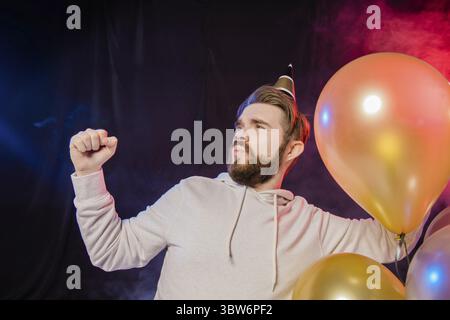 Joyeux anniversaire homme porte en papier chapeau conique danse et chanter et se tient près de ballons d'hélium coloré aime la fête et exprime le bonheur - vacances et Banque D'Images