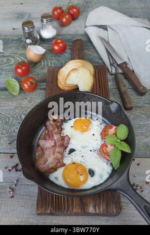 Sur le plat, tranches de bacon, tomate et basilic de moitié dans une poêle en fonte sur une vieille table en bois dans un style rustique Banque D'Images